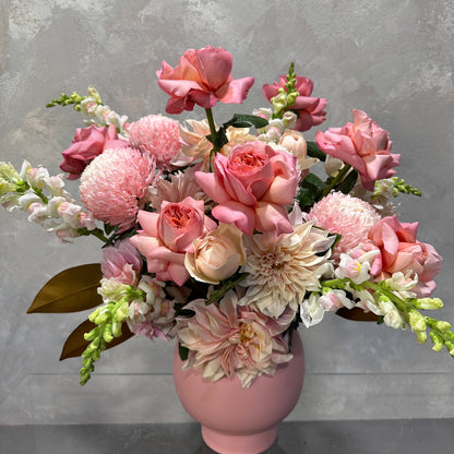 The Bubblegum Pop arrangement from Flowers on Norton St showcases a pink ribbed vase holding a stunning bouquet of pink roses, peonies, and dahlias. This floral display features seasonal blooms, including white and pink snapdragons accented with lush green foliage, all beautifully set against a neutral backdrop.