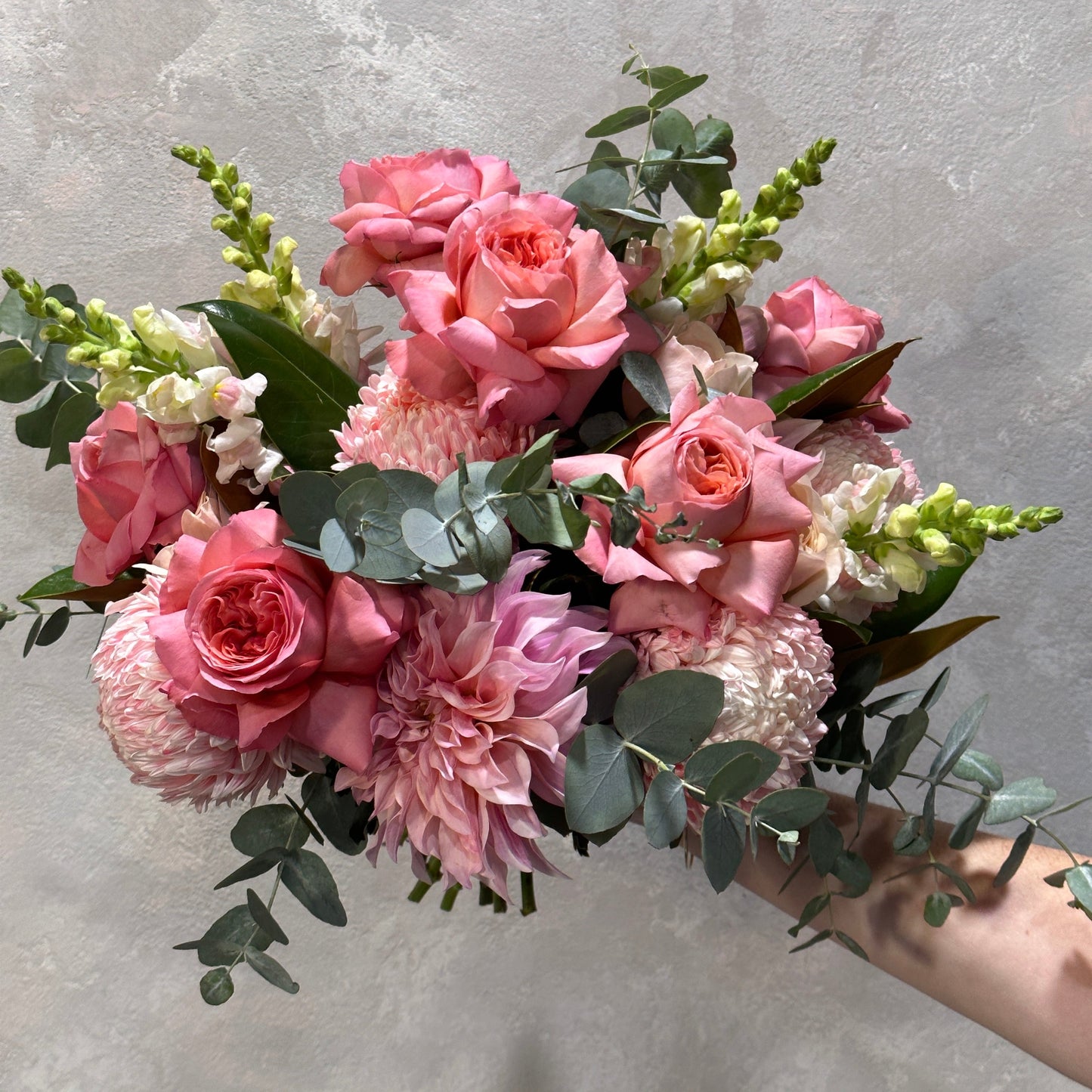 A hand holds the "Pretty in Pink" seasonal bouquet from Flowers on Norton St, showcasing pink roses, dahlias, and snapdragons, accented with silver dollar eucalyptus leaves against a light gray background.