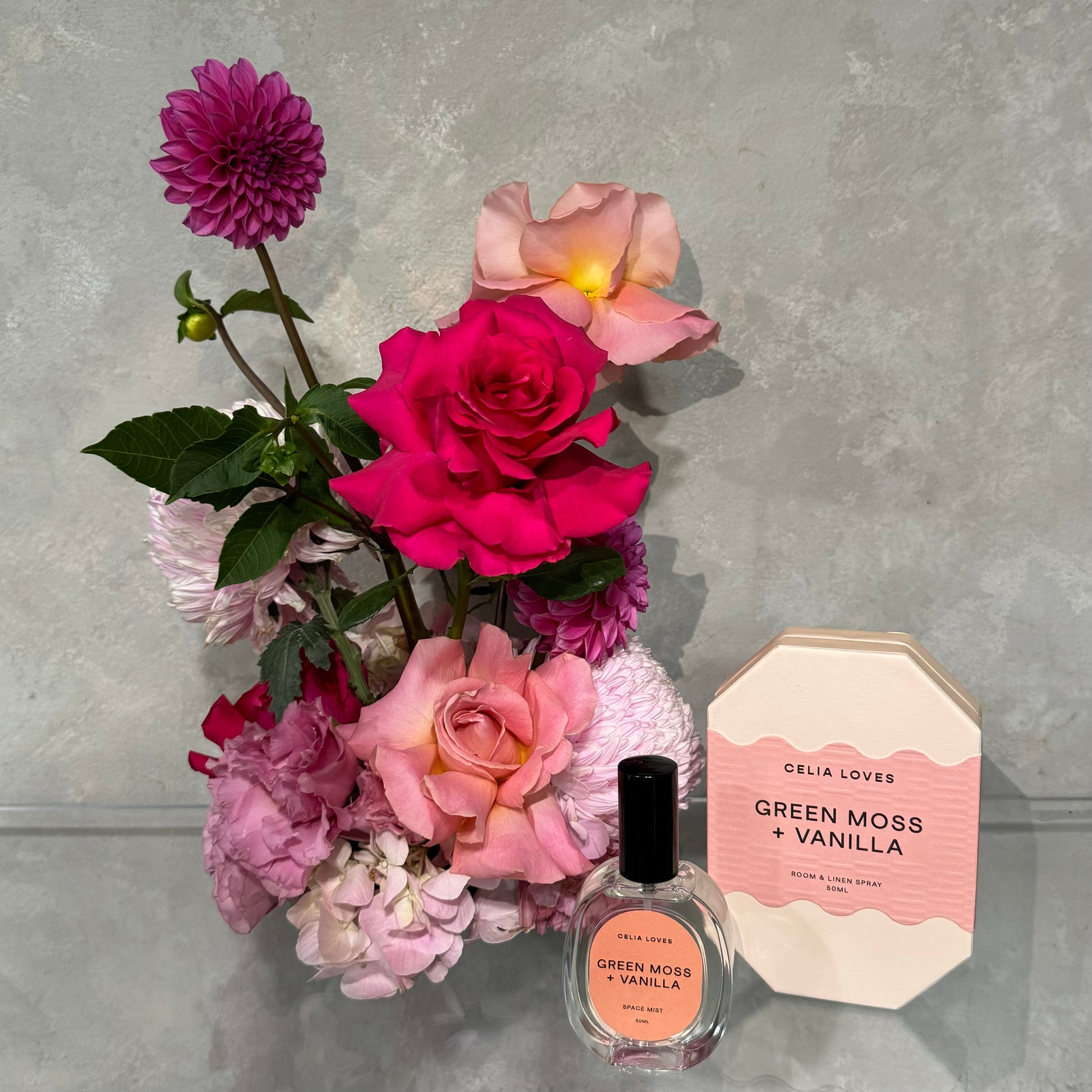 A beautiful floral arrangement featuring pink, purple, and peach blossoms complements the sophisticated "Green Moss + Vanilla" fragrance set. Presented in a beautifully designed package on a gray surface, this ensemble captures the essence of the French Rose Gift Set by Flowers on Norton St.