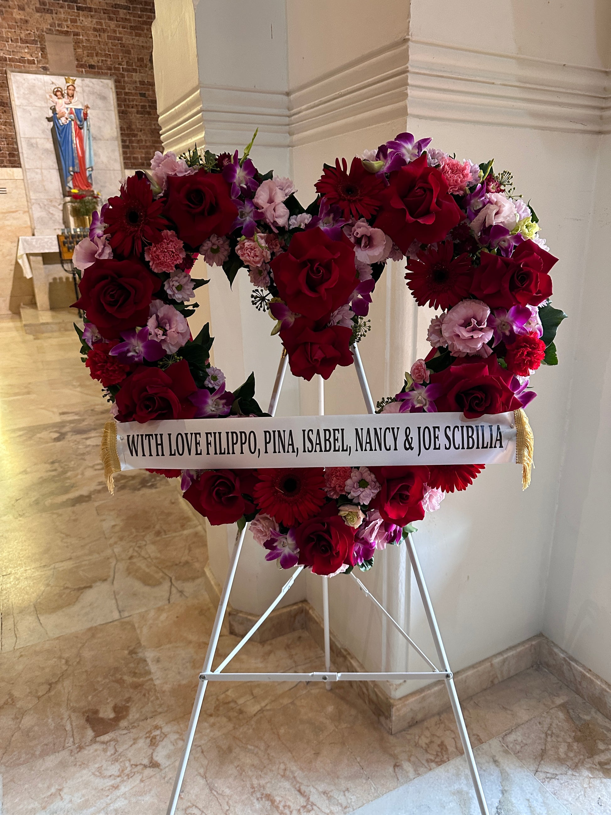 The Heart Wreath Sympathy Flowers by FlowersonNortonSt, crafted from premium red, pink, and purple blooms, is elegantly displayed on a metal easel. A ribbon drapes across the wreath with the message, "With love Filippo, Pina, Isabel, Nancy & Joe Scibilia," while a statue can be seen in the background.