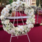 A luxurious circular floral arrangement, featuring the FlowersonNortonSt Large Wreath with white roses, lilies, and orchids, decorates the church's red-carpeted aisle. Displayed on an easel, a ribbon draped across this premium wreath reads, "Sarai un Angelo fra gli angeli Franco, Elisa e famiglia.