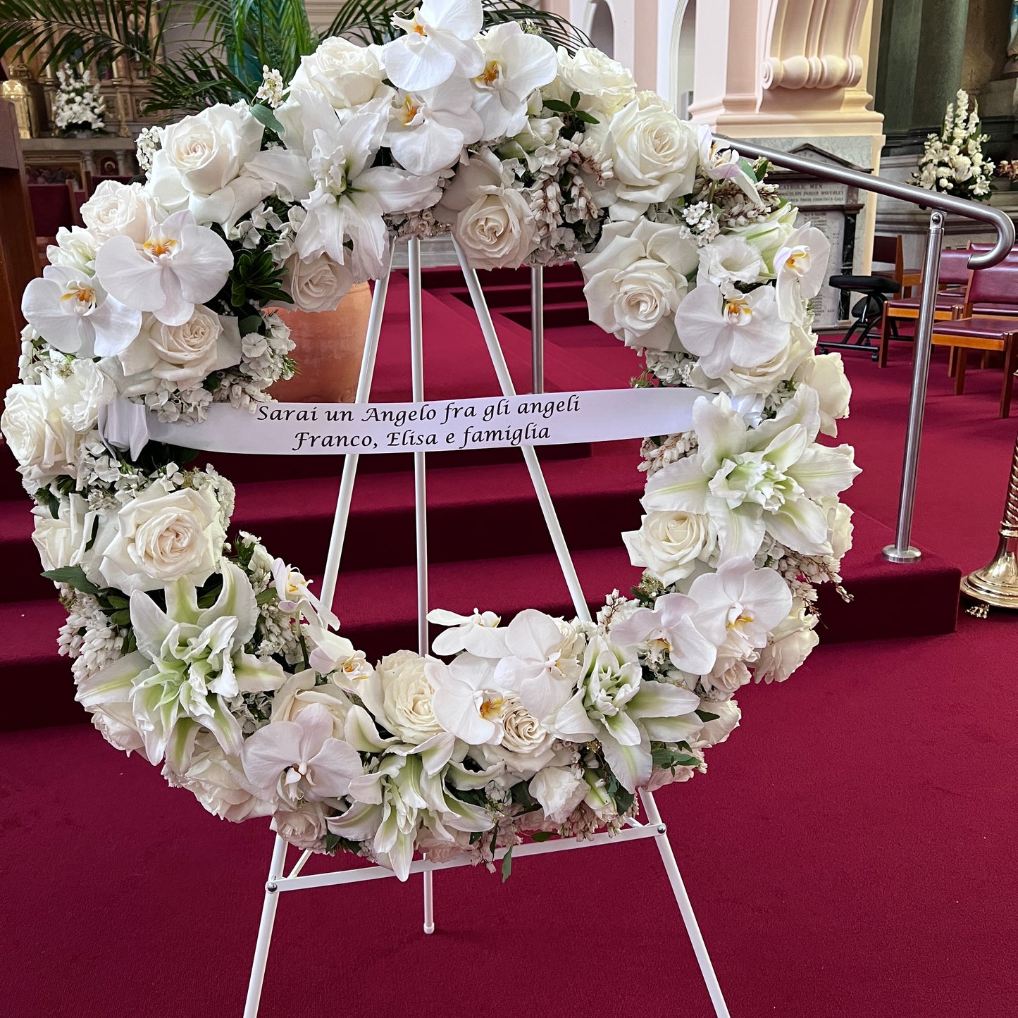 A luxurious circular floral arrangement, featuring the FlowersonNortonSt Large Wreath with white roses, lilies, and orchids, decorates the church's red-carpeted aisle. Displayed on an easel, a ribbon draped across this premium wreath reads, "Sarai un Angelo fra gli angeli Franco, Elisa e famiglia.