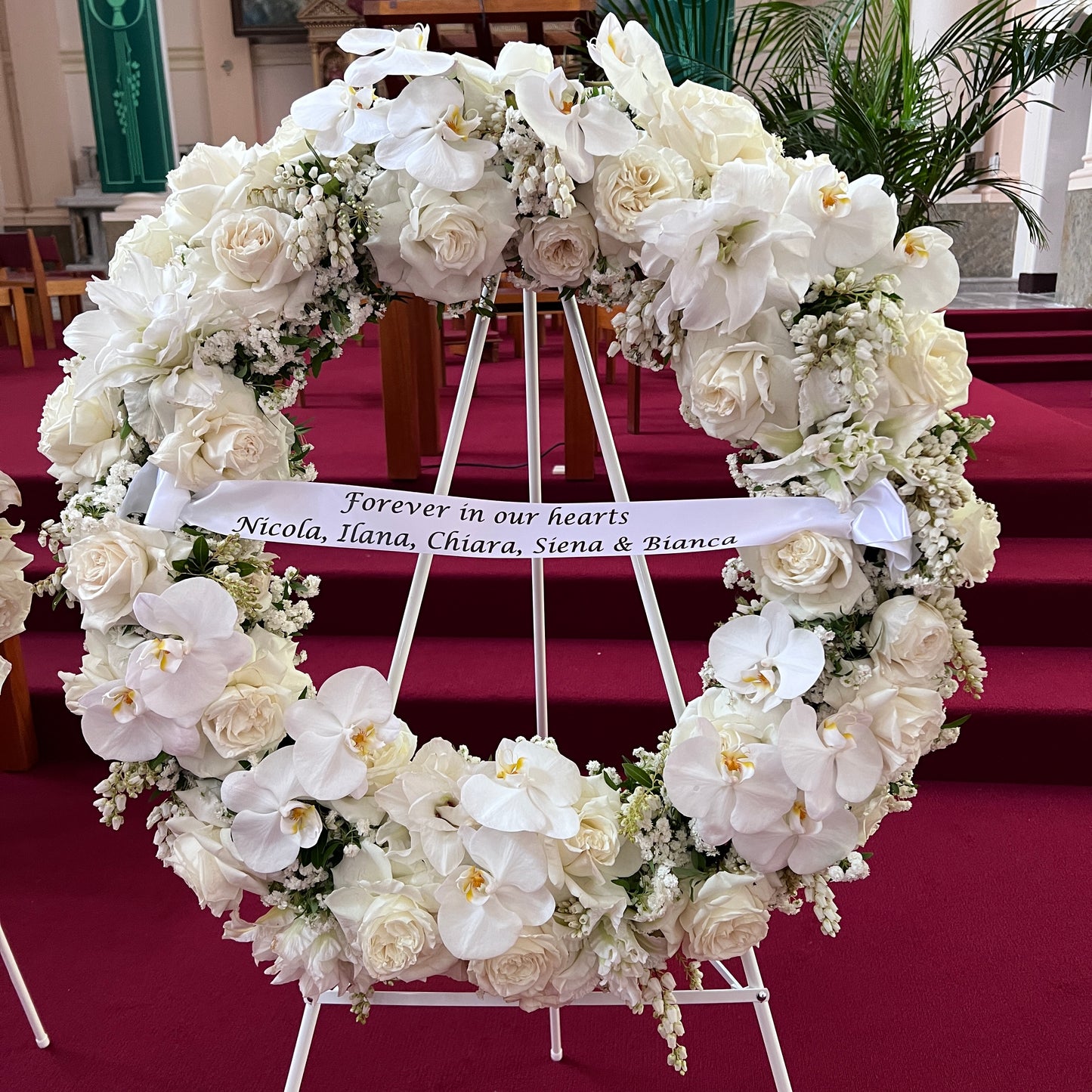 A majestic Large Wreath by FlowersonNortonSt, adorned with luxe roses, orchids, and lilies, stands elegantly in the church. Across this premium wreath is a ribbon inscribed with "Forever in our hearts," along with several names. Red carpeting and green decorations provide a serene backdrop.