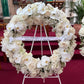 A Large Wreath by FlowersonNortonSt, featuring a premium arrangement of white roses and orchids, is beautifully displayed on an easel in a church setting. A ribbon on the elegant wreath reads, "Forever embedded in our hearts Patricia, Pino & Amilia.