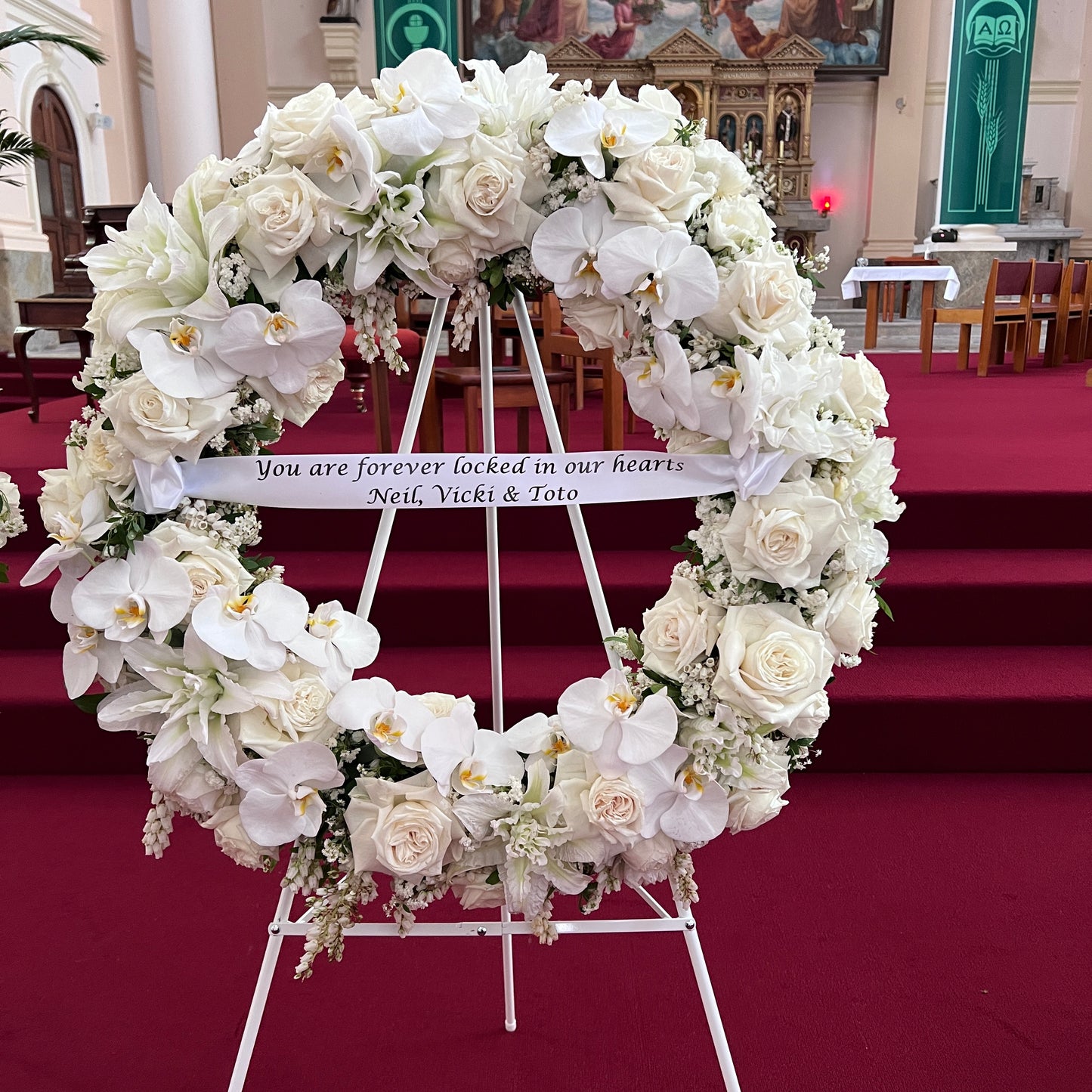 A Large Wreath from FlowersonNortonSt stands elegantly on an easel in a church with red carpeting. Adorned with luxurious white roses and orchids, a ribbon across this premium wreath reads, "You are forever locked in our hearts, Neil, Vicki & Toto.