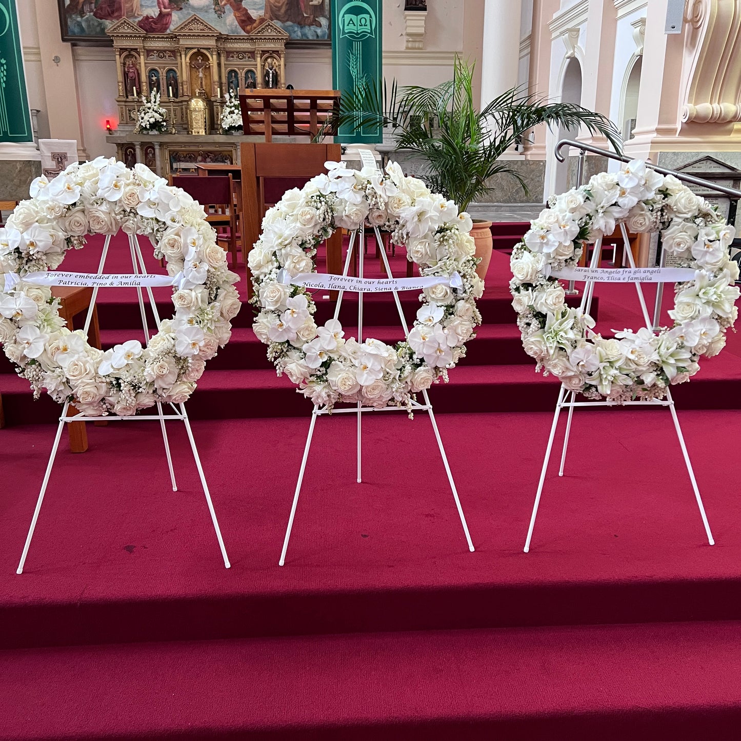 Three large FlowersonNortonSt wreaths are showcased on stands against a red carpet in the church. The ornate interior, adorned with religious artwork and decorative elements, perfectly complements these luxurious Large Wreath arrangements.