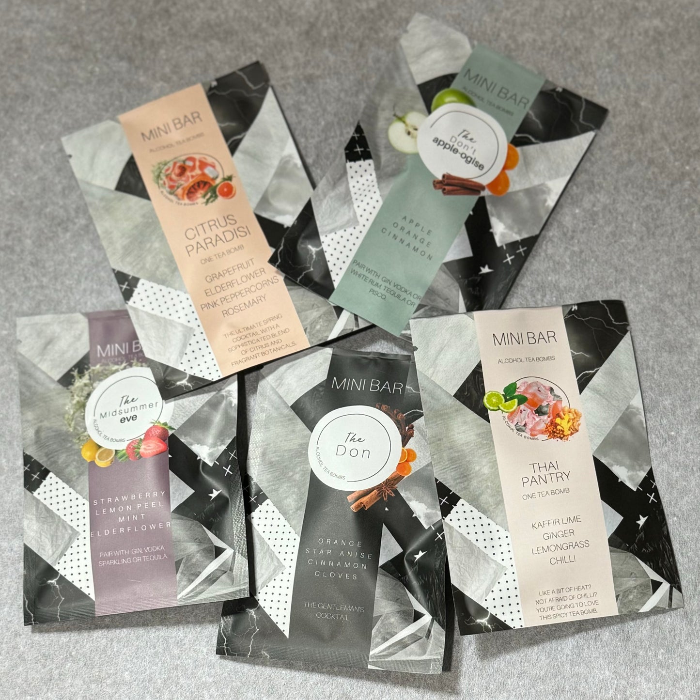 Five vibrant Alcohol Tea-Bomb packets by Flowers on Norton St are displayed on a gray surface. Each packet showcases captivating drink flavor combinations and artistic designs, featuring motifs such as citrus fruits, spices, and herbs. The packets are arranged in a fan-like pattern to highlight all five flavors.