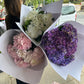 A person holding three Hydrangea Bunches from Flowers on Norton St, lovingly wrapped in white paper and adorned with radiant blooms of white, pink, and purple. Amidst the bustling sidewalk with cars and trees in the backdrop, these vibrant flowers reflect the care of devoted growers.