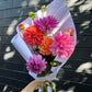 A hand holds a vibrant bouquet of Dahlia Delight wedding flowers from Flowers on Norton St, featuring colorful dahlias in shades of pink, orange, and purple, elegantly wrapped in white paper. The dark brick wall in the background is softly illuminated by dappled sunlight.