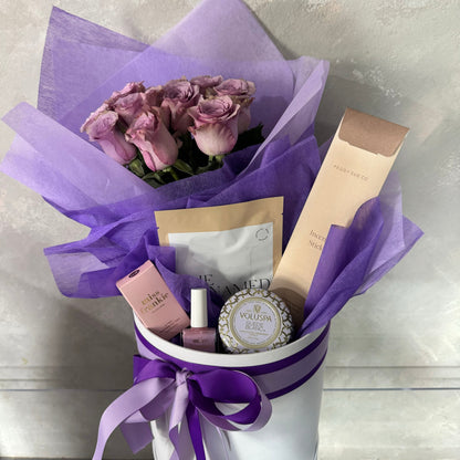 Introducing the Mother's Day Gift Box - Purple Collection by Flowers on Norton St, showcasing exquisite pink roses elegantly wrapped in purple tissue paper. This delightful gift box includes beauty items like a candle, nail polish, and skincare products, all beautifully adorned with a purple ribbon. Available for delivery throughout Sydney.