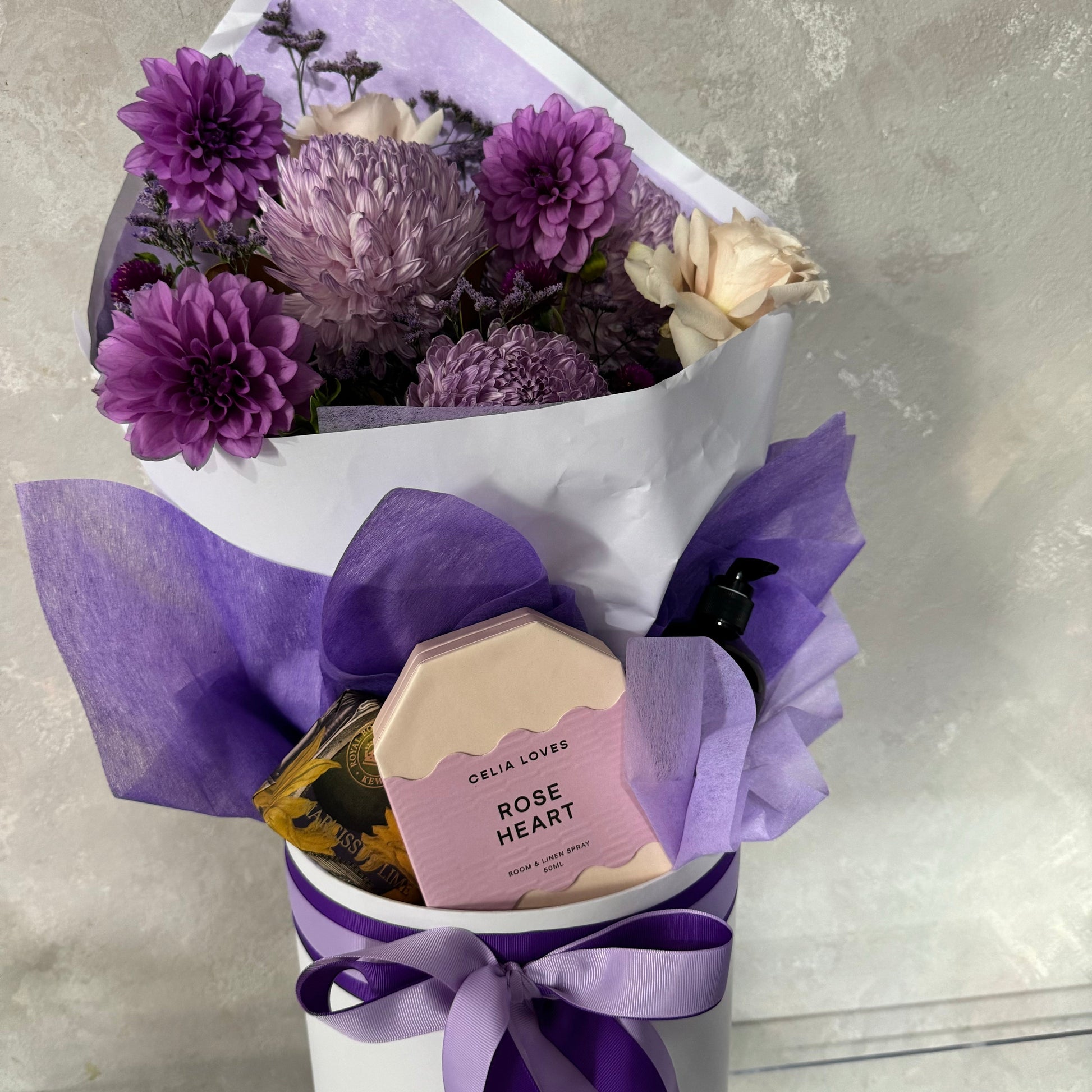 The Mother's Day Gift Box - Purple Collection by Flowers on Norton St is an ideal gift, featuring a bouquet of purple and white flowers in a vase, elegantly wrapped in matching paper with a satin ribbon. This gift set includes a round box labeled "Celia Loves Rose Heart" and a stylish dark pump bottle, all available for Sydney Wide Flower Delivery.