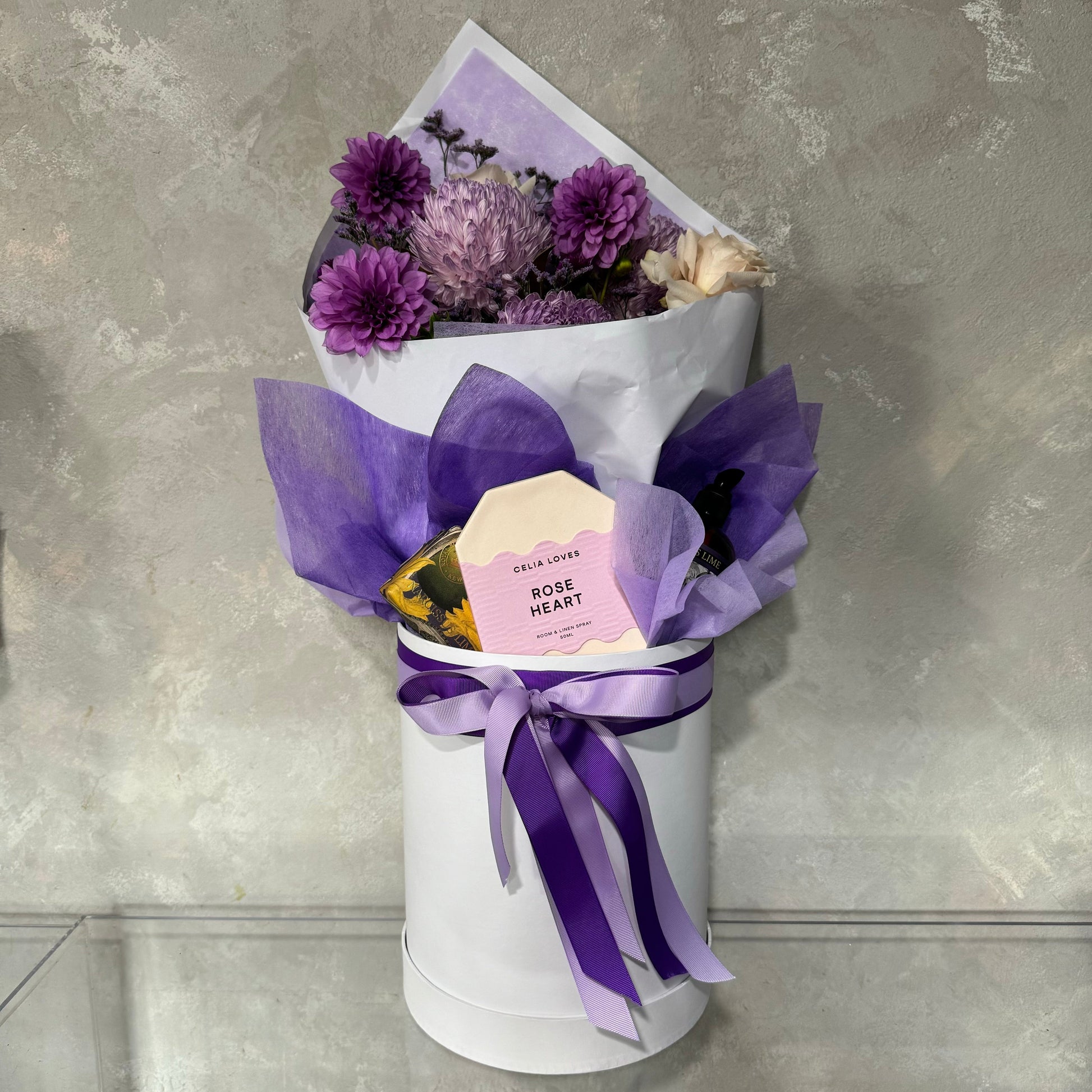 The Mother's Day Gift Box - Purple Collection by Flowers on Norton St features a bouquet of purple flowers elegantly wrapped in white and sheer purple paper, adorned with a purple ribbon, and presented in a round white box. A card is nestled among the blooms. The background is a textured gray wall, perfect for Sydney Wide flower delivery.