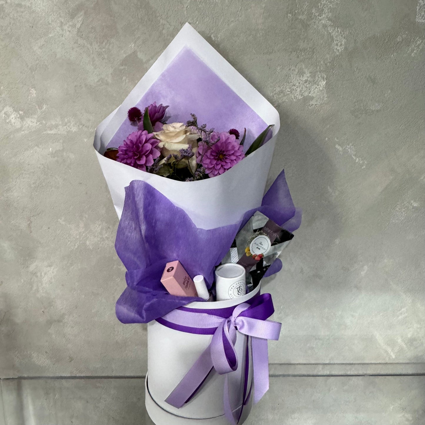 Presenting the Mother's Day Gift Box - Purple Collection by Flowers on Norton St: This exquisite arrangement boasts a stunning combination of purple and white flowers, elegantly wrapped in matching paper. The bouquet is beautifully displayed in a white cylindrical base, adorned with a purple ribbon. This gift set is ideal for Mother's Day, featuring delightful surprises such as a pink box and a white canister—perfect for flower delivery across Sydney.