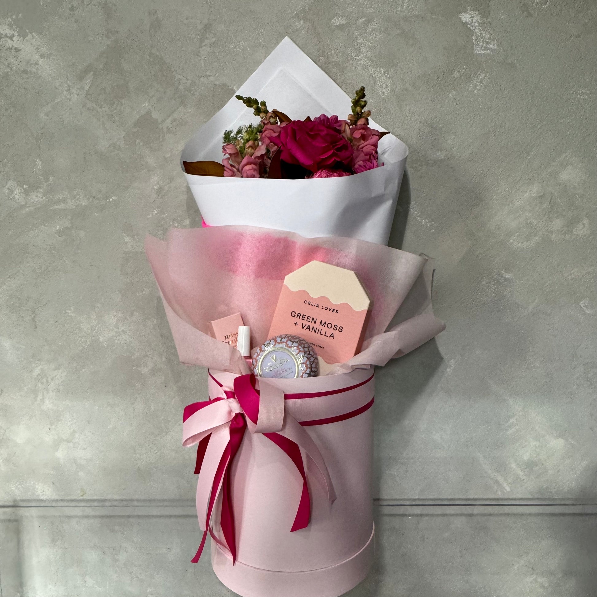 A bouquet from Flowers on Norton St's Mother's Day Gift Box - Pink Collection, showcasing pink and red flowers encased in delicate paper, is nestled in a pink cylindrical box with a ribbon. A label displaying "Green Moss & Vanilla" completes this elegant arrangement, set against a gray textured wall for the ultimate pampering experience.