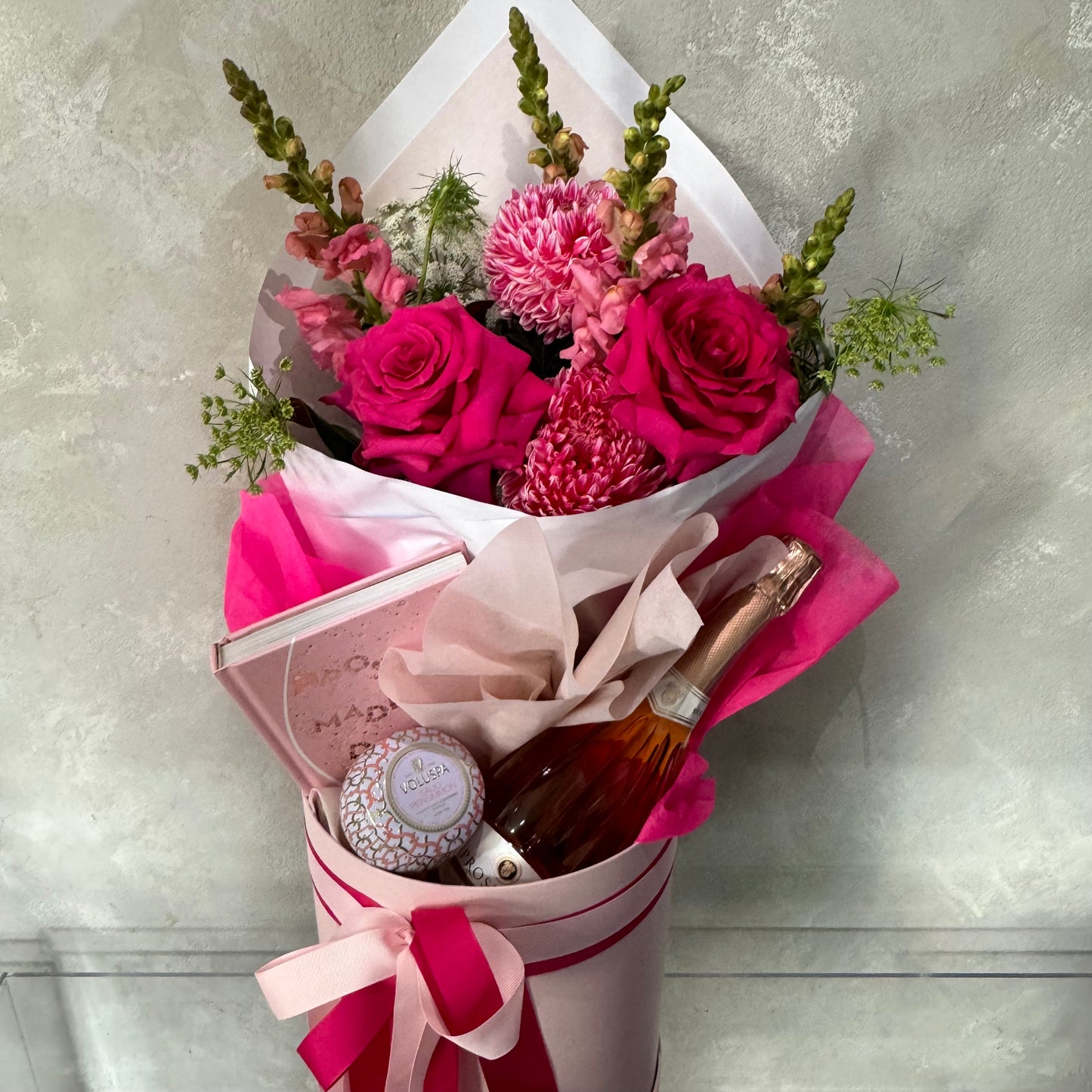 The Mother's Day Gift Box - Pink Collection from Flowers on Norton St features a delightful bouquet of pink roses and flowers, paired with a bottle of rosé, a pink book, and a small jar of cream. Elegantly presented in a pink box adorned with a decorative bow, it offers the ultimate pampering experience.