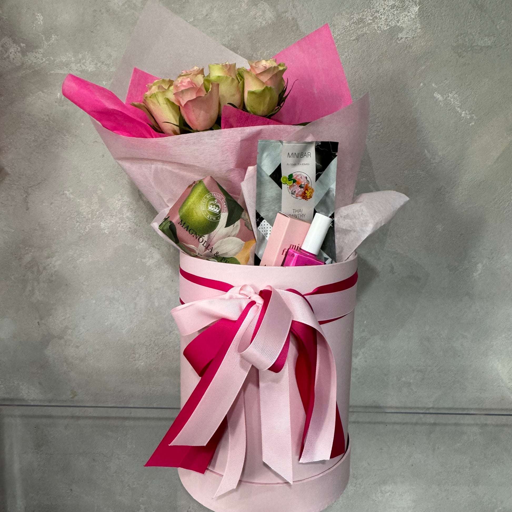 The Mother's Day Gift Box - Pink Collection by Flowers on Norton St offers a luxurious pampering experience, adorned with a large pink and white ribbon that enwraps stunning pink roses, elegantly wrapped in sophisticated paper. It also includes an exquisite selection of cosmetic products against an elegant gray backdrop.