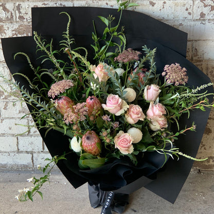 The "Delicate Roses + Natives" bouquet by Flowers on Norton St, showcasing delicate roses, protea flowers, and a selection of greenery, is elegantly wrapped in black paper and displayed against a rustic brick wall.