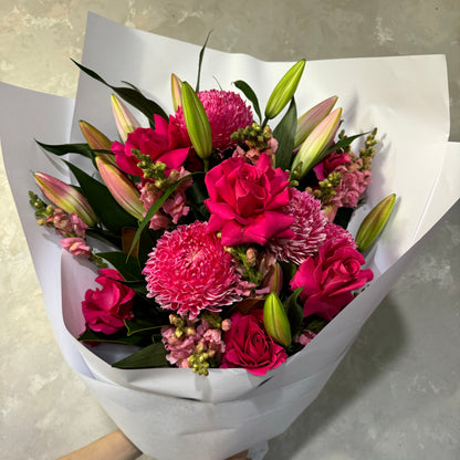 The "Watermelon Crush" by Flowers on Norton St is an exquisite Mother's Day bouquet of vibrant pink blooms, including roses, chrysanthemums, and Oriental lilies, elegantly wrapped in white paper. It's beautifully arranged against a textured gray backdrop.