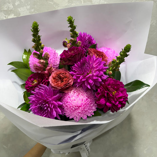 The Floral Fusion Bouquet for Mum by Flowers on Norton St is a vibrant mix of purple, pink, and red flowers. This elegant gift for mum features dahlias, roses, lush green leaves and other bright blooms wrapped in white paper to create a colorful display.