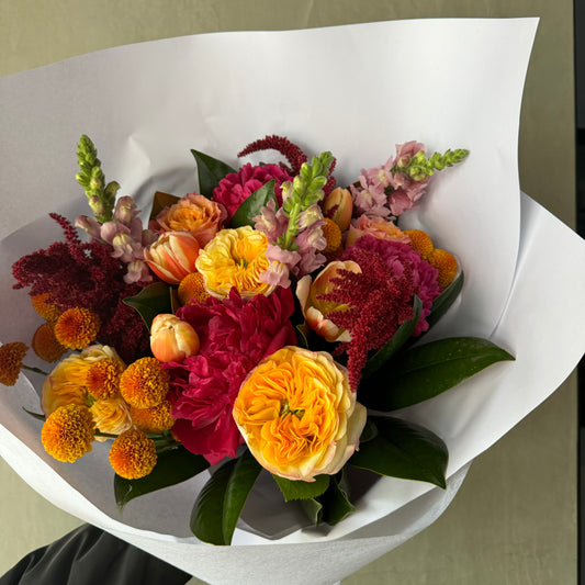 The Spring Sunrise Bouquet from Flowers on Norton St offers a vibrant mix of yellow roses, pink peonies, orange ranunculus, red amaranthus, pink snapdragons, and small orange chrysanthemums wrapped in crisp white paper with lush green leaves.