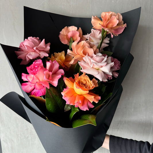 A person elegantly holds the "Statement Roses Bouquet 2024" by Flowers on Norton St, featuring hand-crafted Colombian roses in pink, peach, and orange hues wrapped in black paper against a gray background.