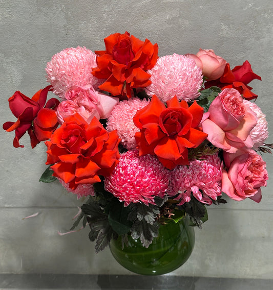 The Strawberry Pinks + Reds Vase Arrangement from Flowers on Norton St features vibrant red and pink roses with pink pompom chrysanthemums, all beautifully displayed in a green vase against a neutral backdrop.