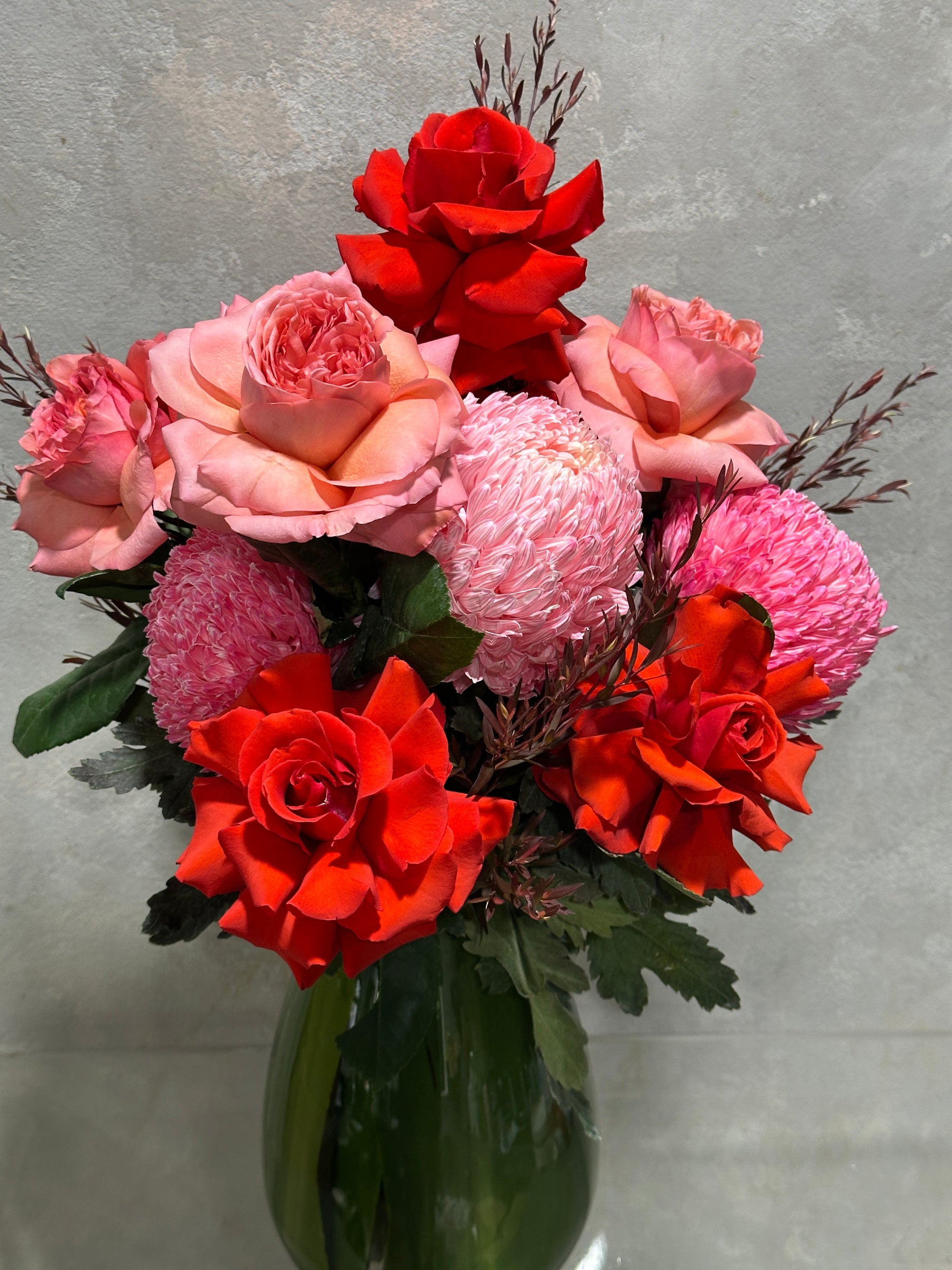 The Strawberry Pinks + Reds Vase Arrangement from Flowers on Norton St features vibrant red and pink flowers, with elegant roses and delicate chrysanthemums in a green vase against a neutral backdrop.