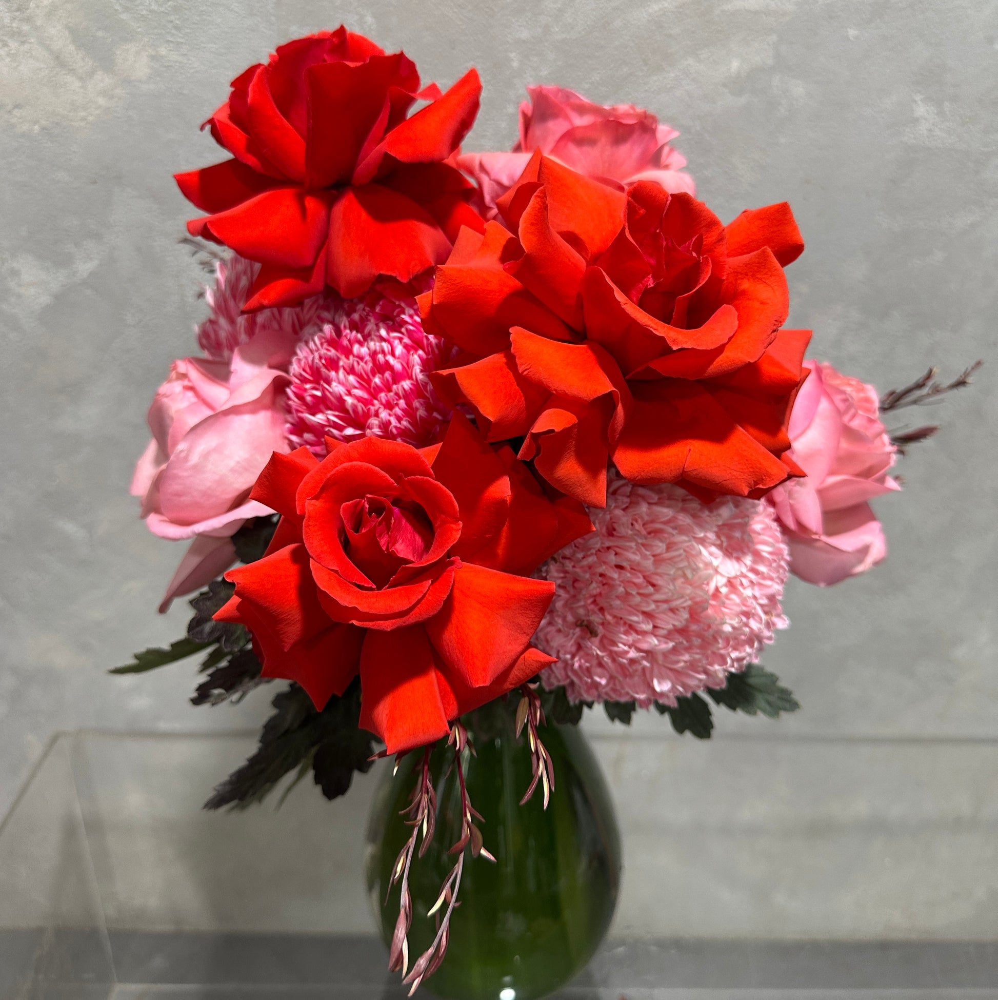 The Strawberry Pinks + Reds Vase Arrangement by Flowers on Norton St features lush red and pink roses and chrysanthemums in a chic green vase against a textured gray backdrop, highlighting the vibrant blooms.