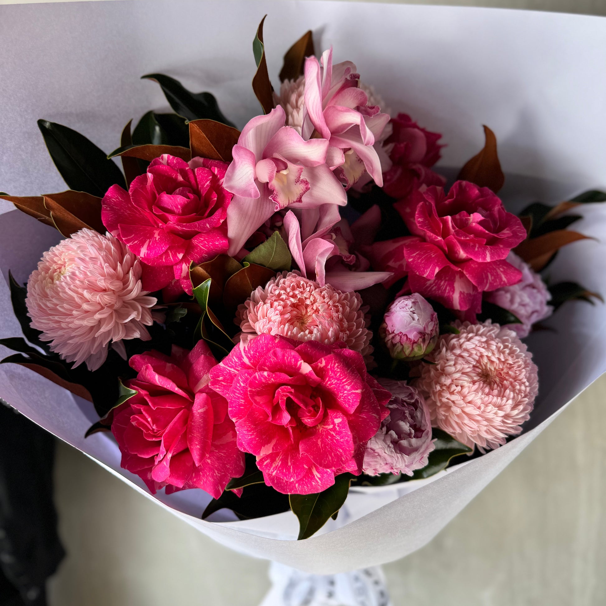 The Raspberry Ripple Bouquet from Flowers on Norton St features a stunning arrangement of vibrant pink flowers like Pandora roses and peonies, combined with rich dark green leaves, all elegantly wrapped in pristine white paper.