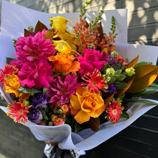 The "Big Bouquet of Flowers" by Flowers on Norton St is a lively arrangement of pink roses, orange gerberas, yellow roses, purple lisianthus, and dahlias. It comes beautifully wrapped in white paper with greenery, set against a textured brick wall background.