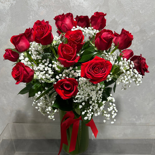 The Classical Lovers Red Rose Vase by Flowers on Norton St features romantic red roses and delicate baby's breath, elegantly arranged with a red ribbon, set against a plain gray background.