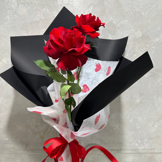 The "Just the Two of Us Roses" by Flowers on Norton St features two vibrant Colombian red roses in black wrapping with red hearts and a romantic red ribbon, making it an ideal Valentine's Day gift against a light textured background.