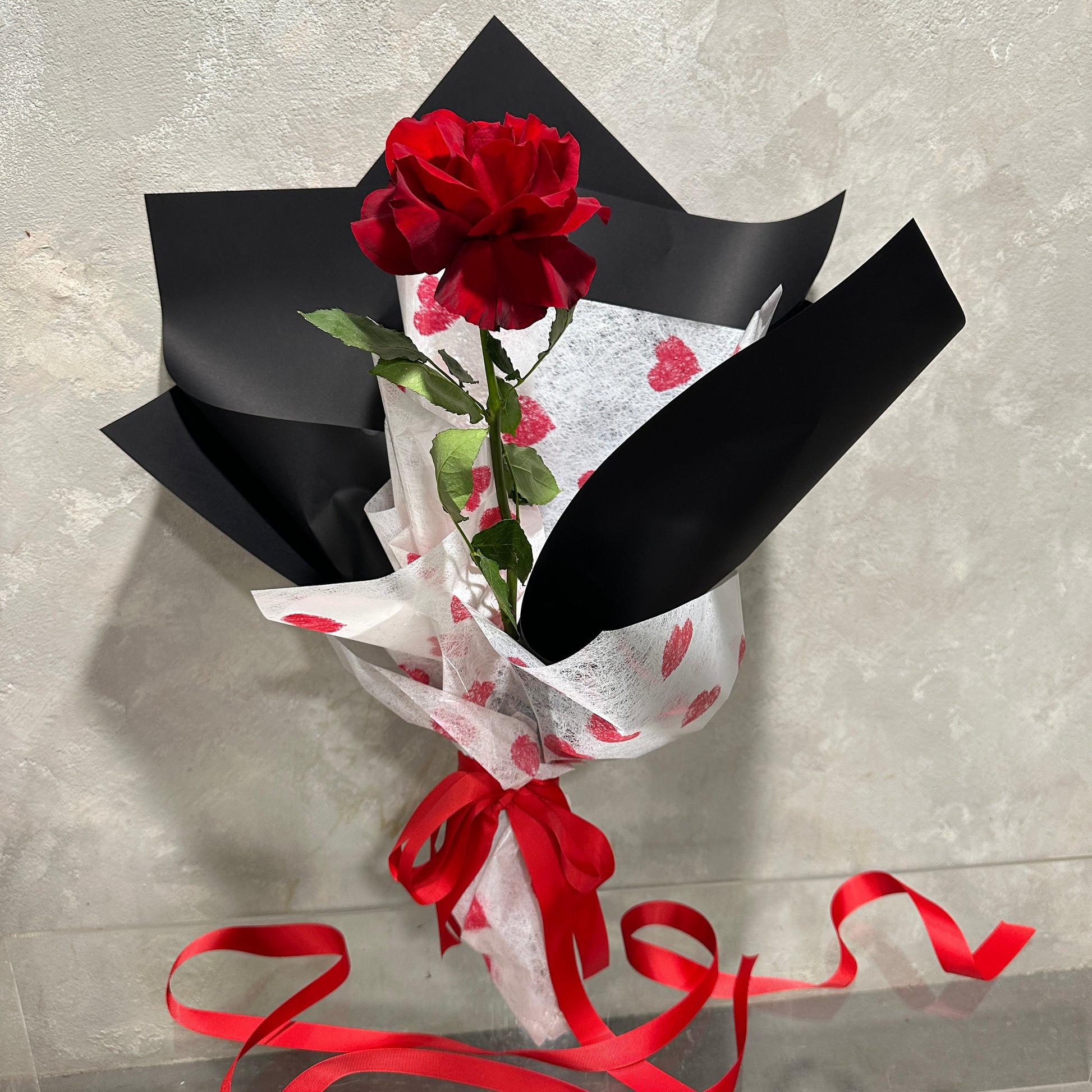 The "My One and Only Red Rose" by Flowers on Norton St features a single red rose wrapped in heart-patterned white paper with black accents, tied with a red ribbon. This charming bouquet, perfect for Valentine's Day, is set against a textured gray background.