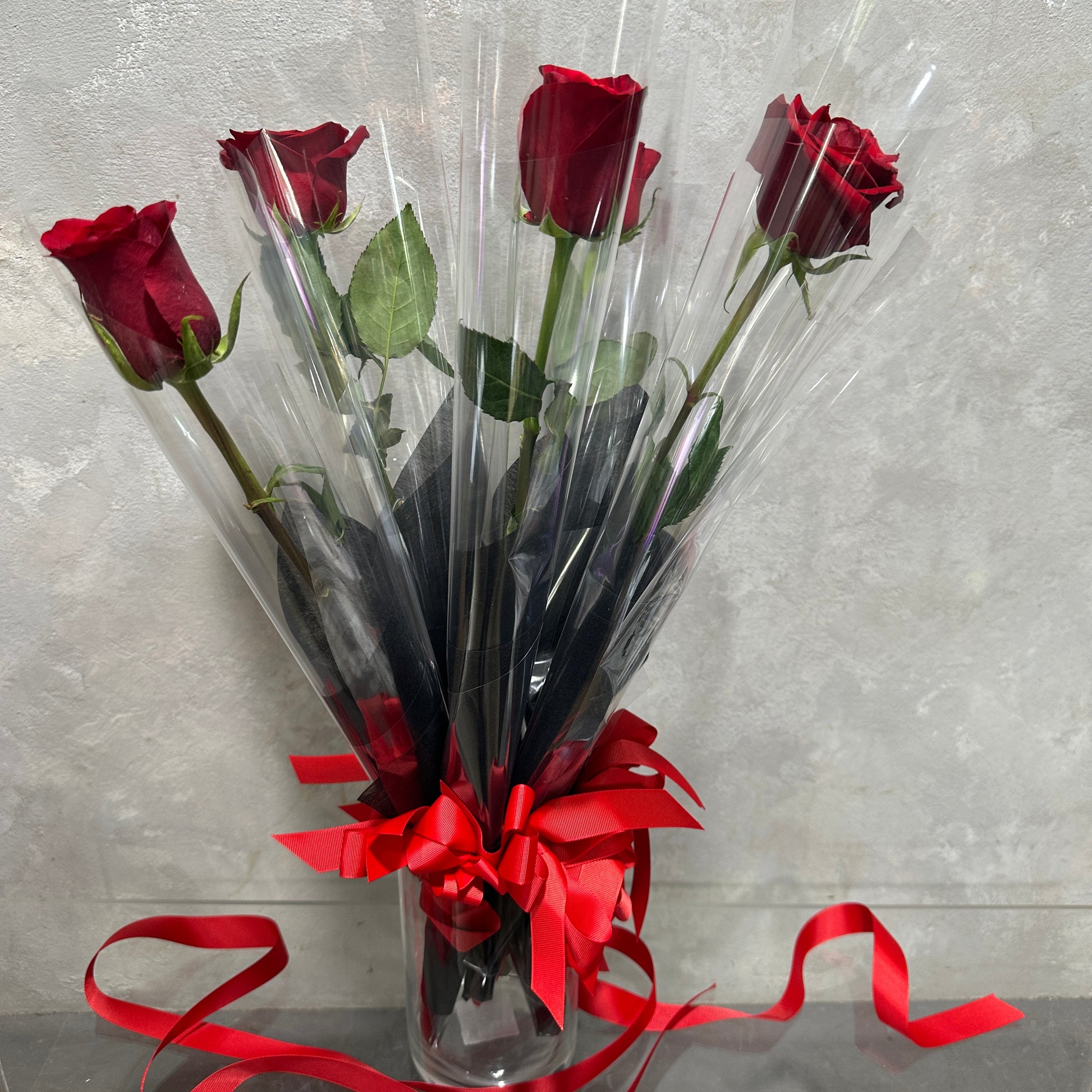 The "Single Red Rose" from Flowers on Norton St is elegantly wrapped in clear plastic with a vibrant red ribbon, displayed in a tall vase against a light gray background, adding timeless charm to any setting.