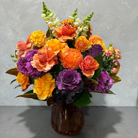 A vibrant floral arrangement, presented in the "Amber Tulip Vase" from Flowers on Norton St, showcases hand-flexed orange and peach roses, purple carnations, marigolds, snapdragons, and decorative foliage against a textured gray background.