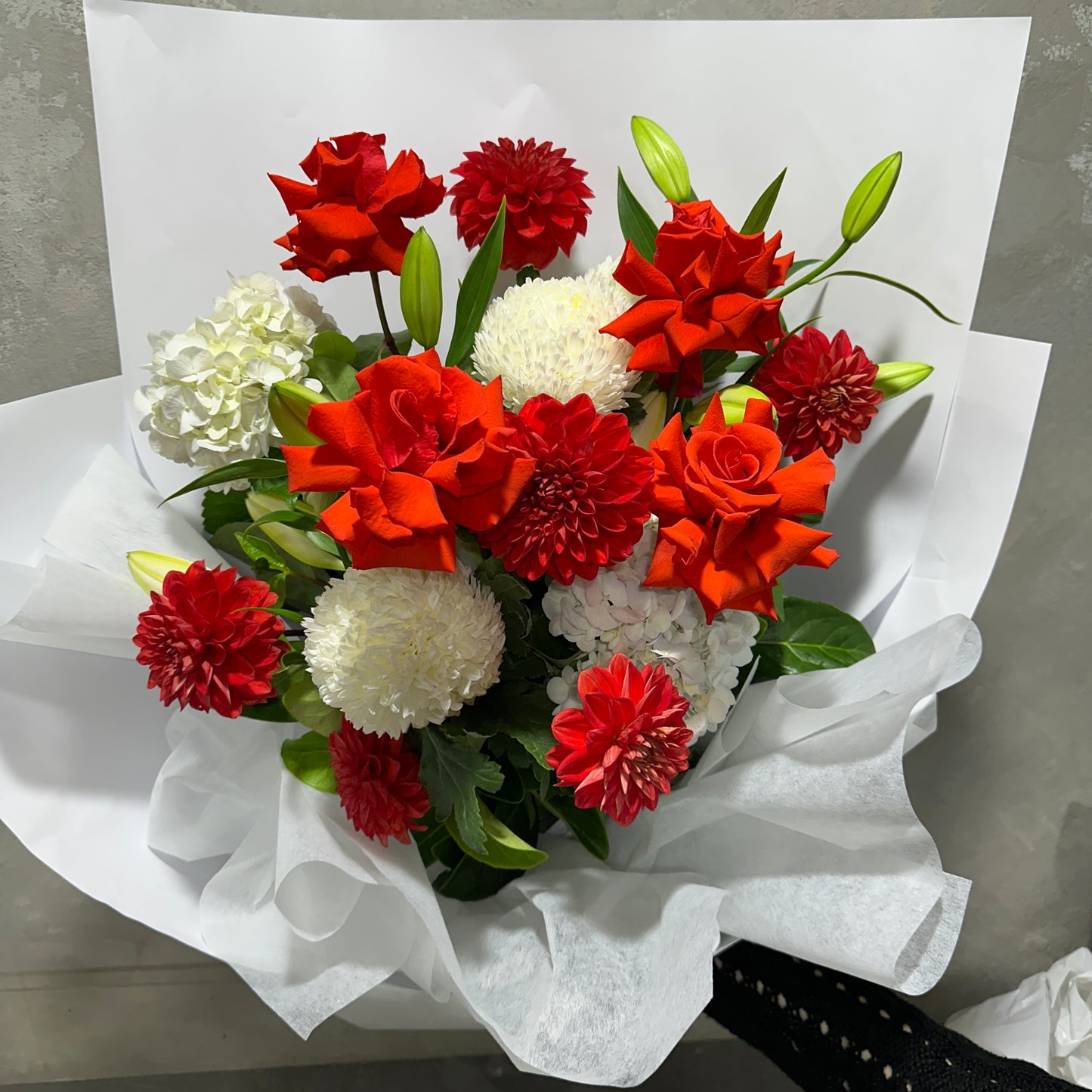 The Red Nina Expression Bouquet by Flowers on Norton St features a vibrant mix of red and white flowers, including roses, dahlias, and chrysanthemums, with green leaves. Wrapped in white paper, it creates a striking contrast against the colorful blooms.