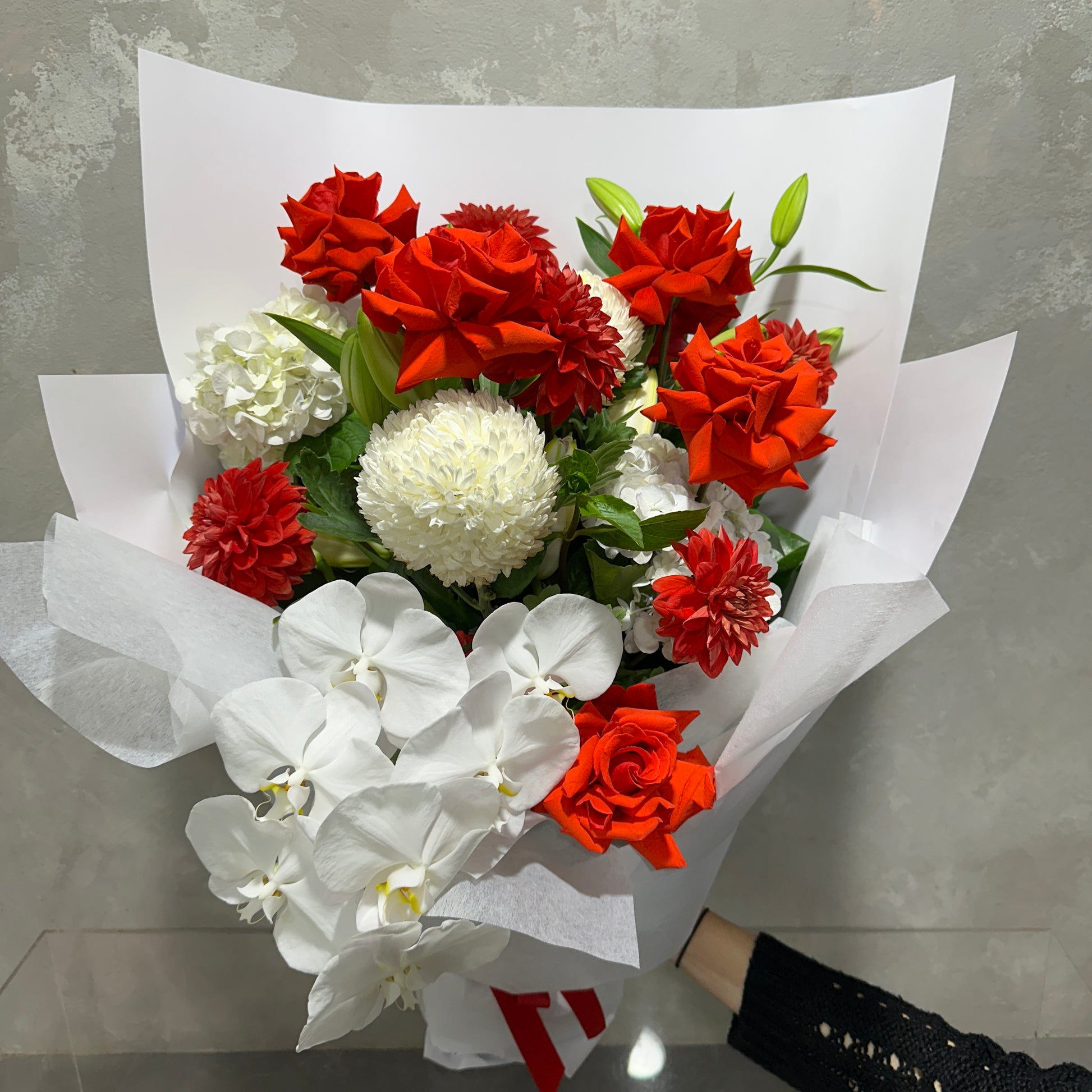 The Red Nina Expression Bouquet by Flowers on Norton St features red roses, dahlias, white hydrangeas, and orchids wrapped in white paper. It's held by a person in a black sweater against a light grey textured background.