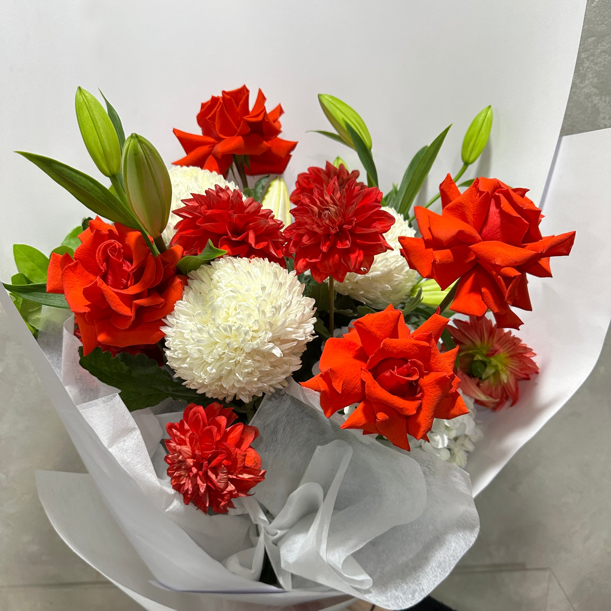 The Red Nina Expression Bouquet from Flowers on Norton St includes vibrant, fresh red roses, red chrysanthemums, white carnations, and white chrysanthemums. It's elegantly wrapped in translucent white paper with green leaves visible.