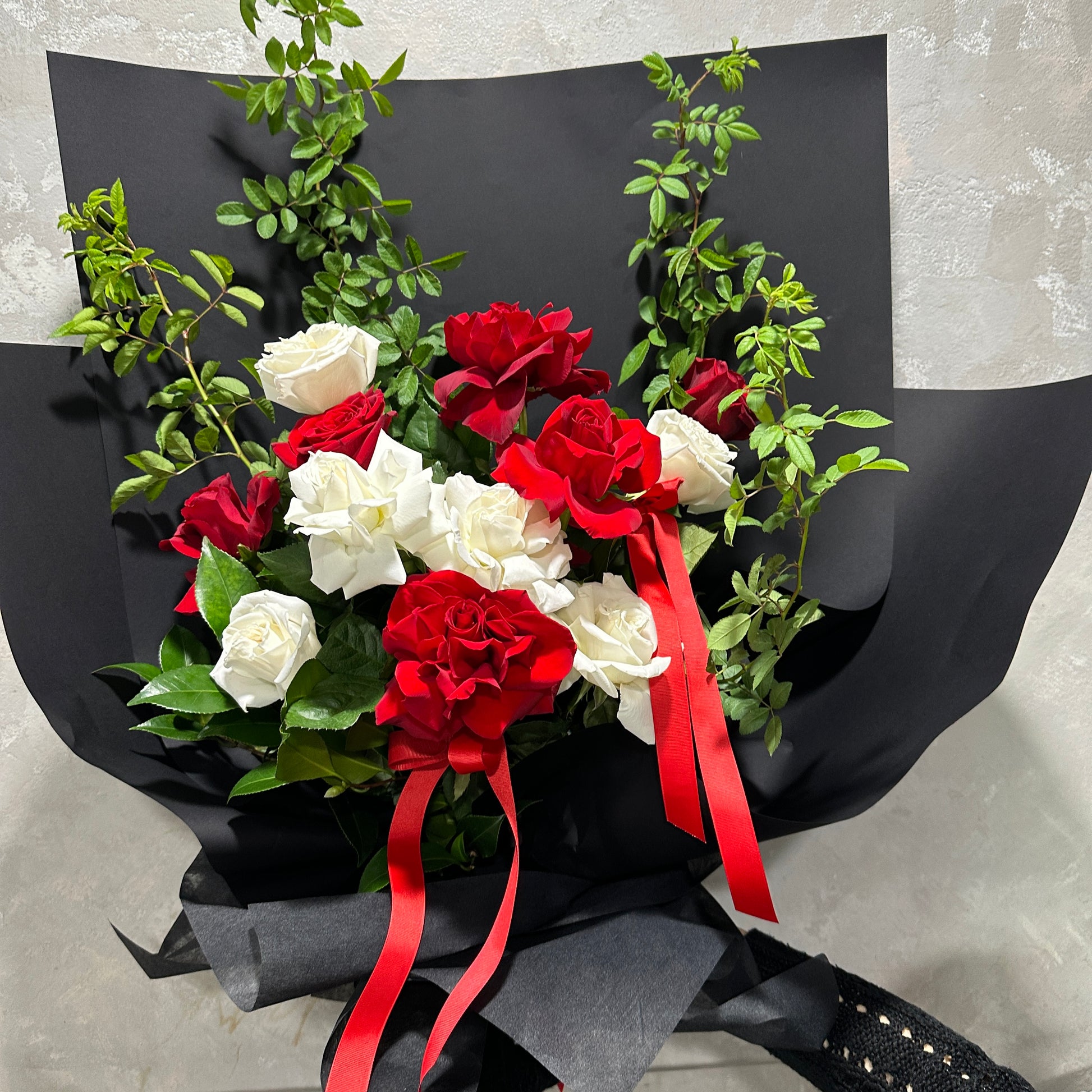 Introducing the Flowers on Norton St Red and White Bud + Bloom Rose Bouquet: a stunning arrangement of 12 red and white roses, elegantly accented with greenery. Wrapped in sleek black paper, it's completed with sophisticated red ribbons around the stems.