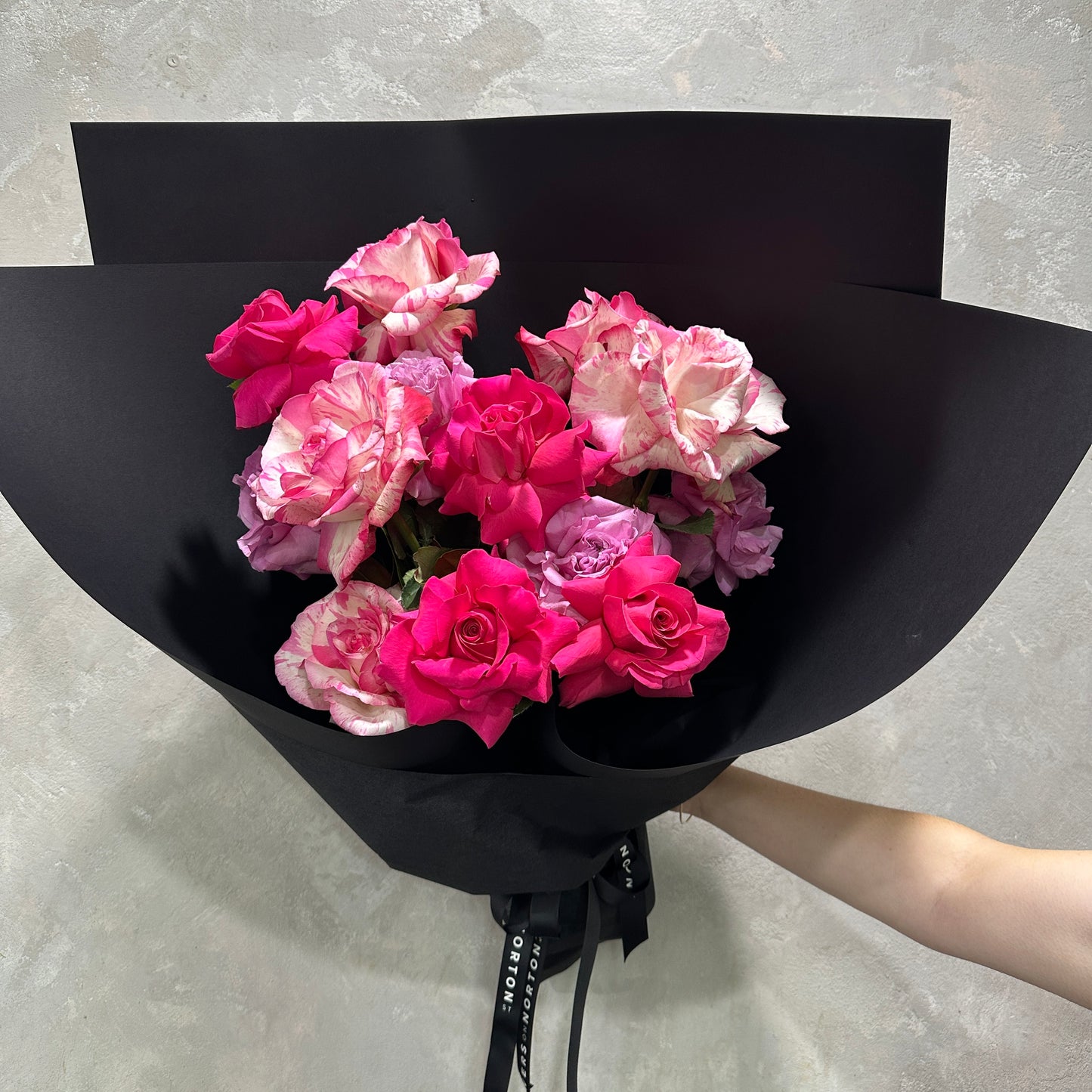 The Rose Trio Bouquet by FlowersonNortonSt features a mix of pink and white handcrafted Colombian roses, beautifully gift wrapped in black paper with a textured light gray background.