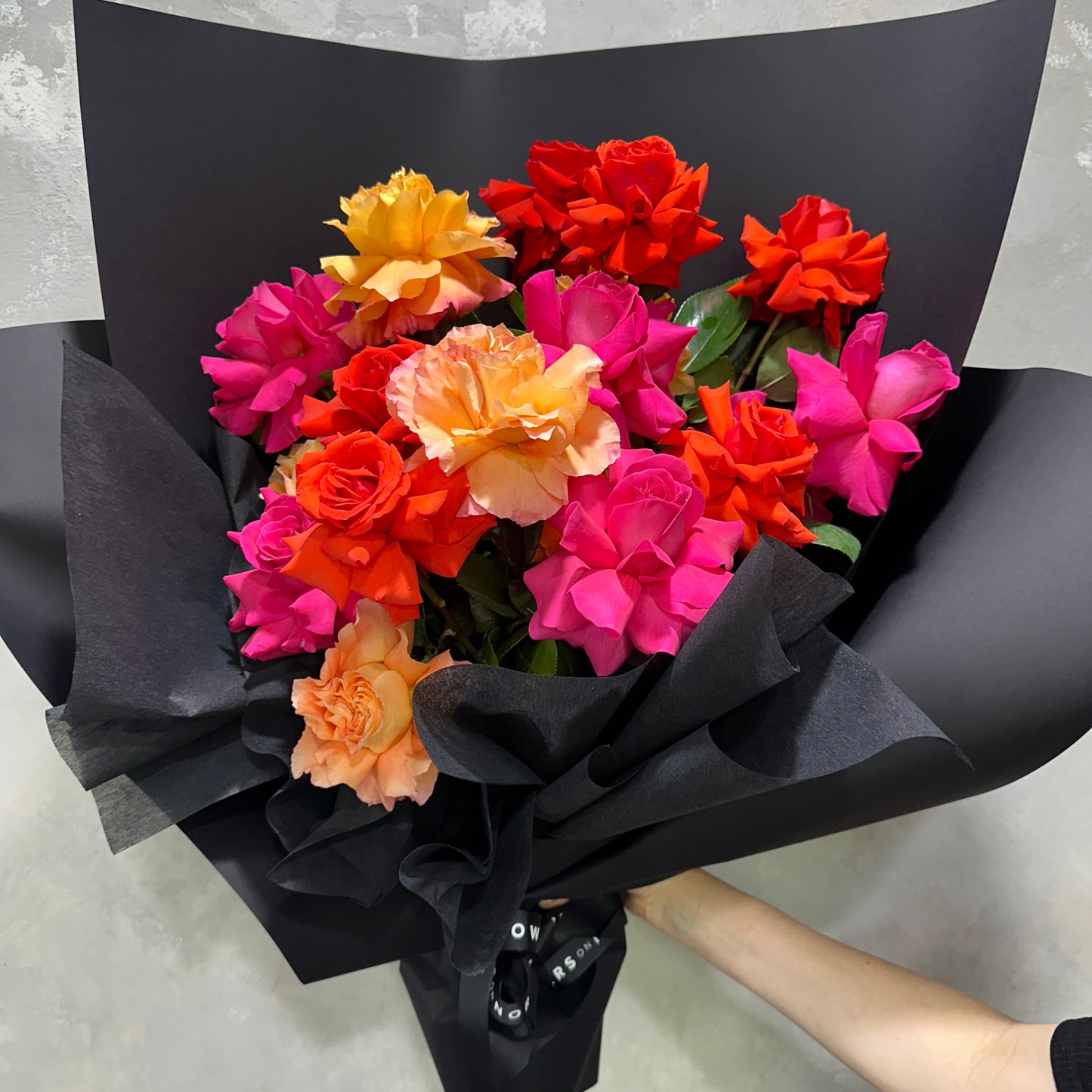 The Rose Trio Bouquet from FlowersonNortonSt features a vibrant mix of handcrafted Colombian roses in red, orange, pink, and yellow. Elegantly wrapped in black paper, this exquisite arrangement is held against a textured gray background.