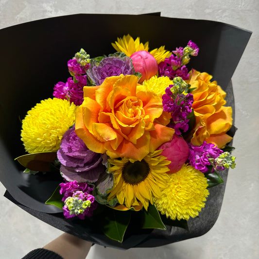 The Lemony Goodness | June Mixed Bouquet by Flowers on Norton St showcases a vibrant mix of orange roses, seasonal foliage, sunflowers, pink peonies, and purple flowers in black paper, creating a striking contrast against any neutral background.