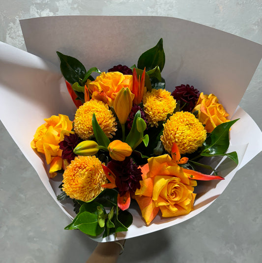 The "Mandarin | June Mixed Bouquet" by Flowers on Norton St features vibrant yellow and orange roses, yellow marigolds, dark red blooms, and delicate lilies wrapped in white paper with green leaves on a textured light gray background.