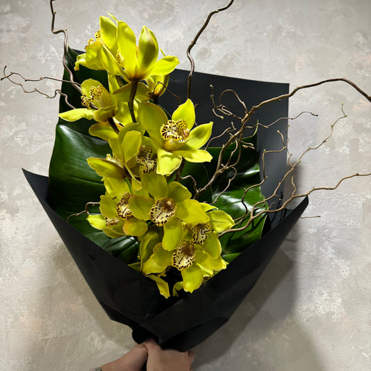 Introducing the Sculptural Cymbidium Bouquet from Flowers on Norton St, showcasing vibrant yellow orchids with spotted centers, complemented by curly branches and lush green leaves, all elegantly wrapped in black paper for a modern touch.