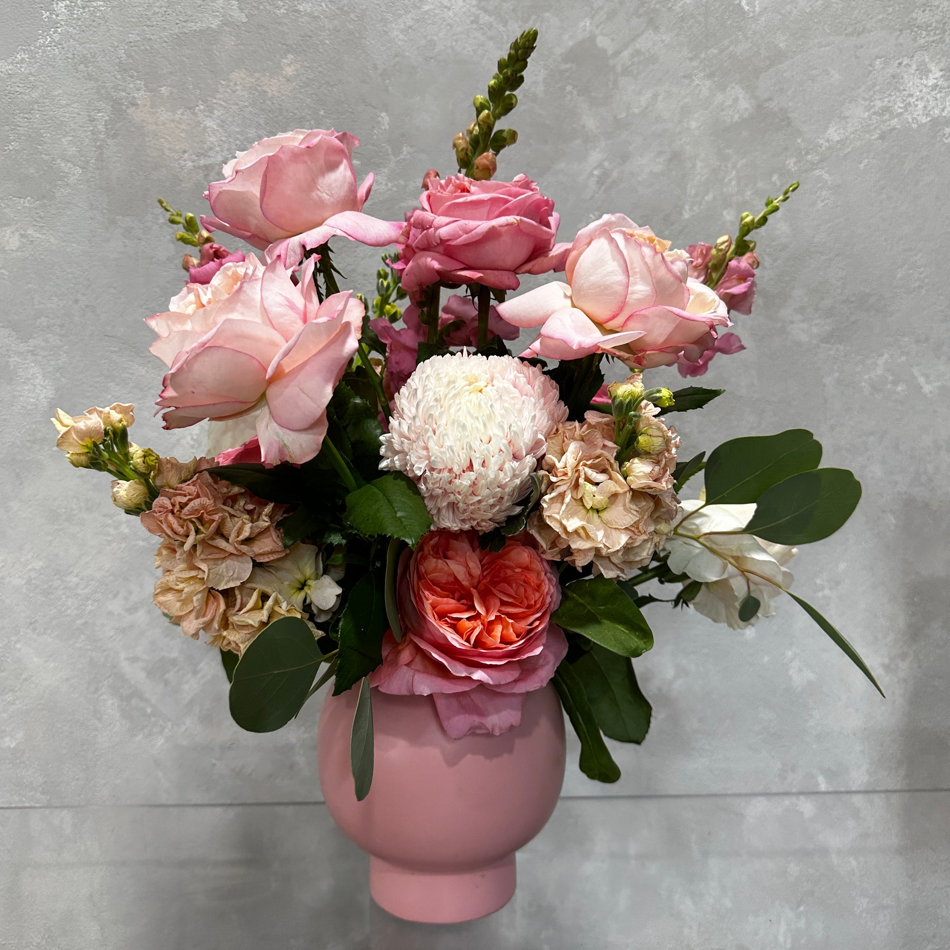 The "Bubblegum Pop" by Flowers on Norton St features a delightful floral arrangement of pink and peach roses, white carnation-like flowers, lush green leaves, and other small blossoms elegantly displayed in a pink ribbed vase against a textured gray background.