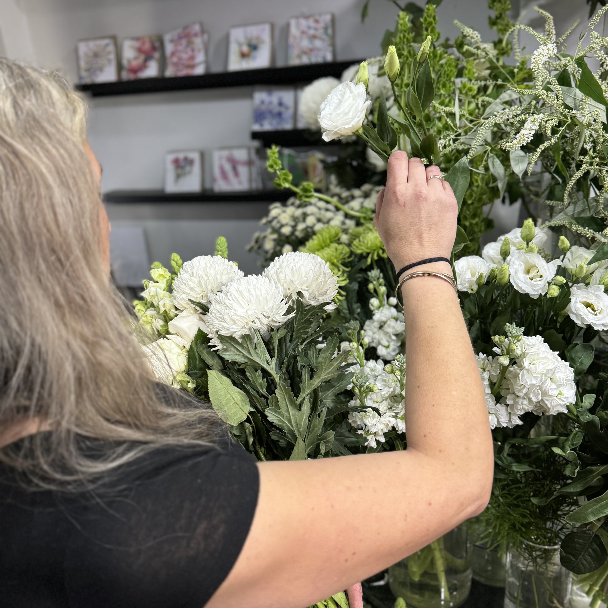 In a bright floral shop, a person with long gray hair arranges a Florist Choice - White bouquet from Flowers on Norton St amid shelves of vibrant white and green blooms. Wearing a black shirt, they craft beauty with care, capturing the elegance offered by our affordable flower delivery Sydney service.