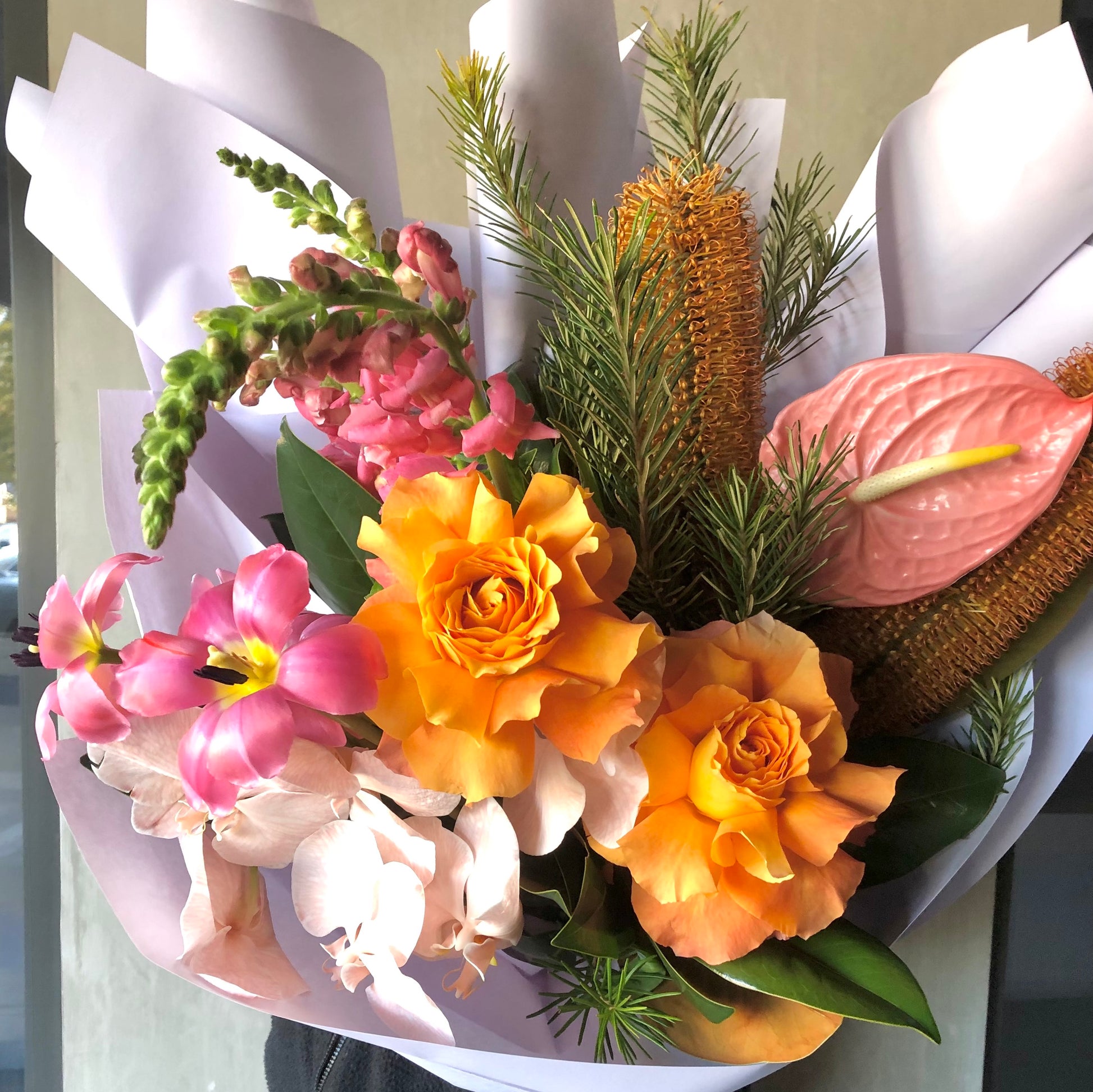 Introducing "Candlelight" by Flowers on Norton St, a stunning floral arrangement of orange roses, pink snapdragons, anthuriums, tropical blooms, and lush greenery elegantly wrapped in white paper.