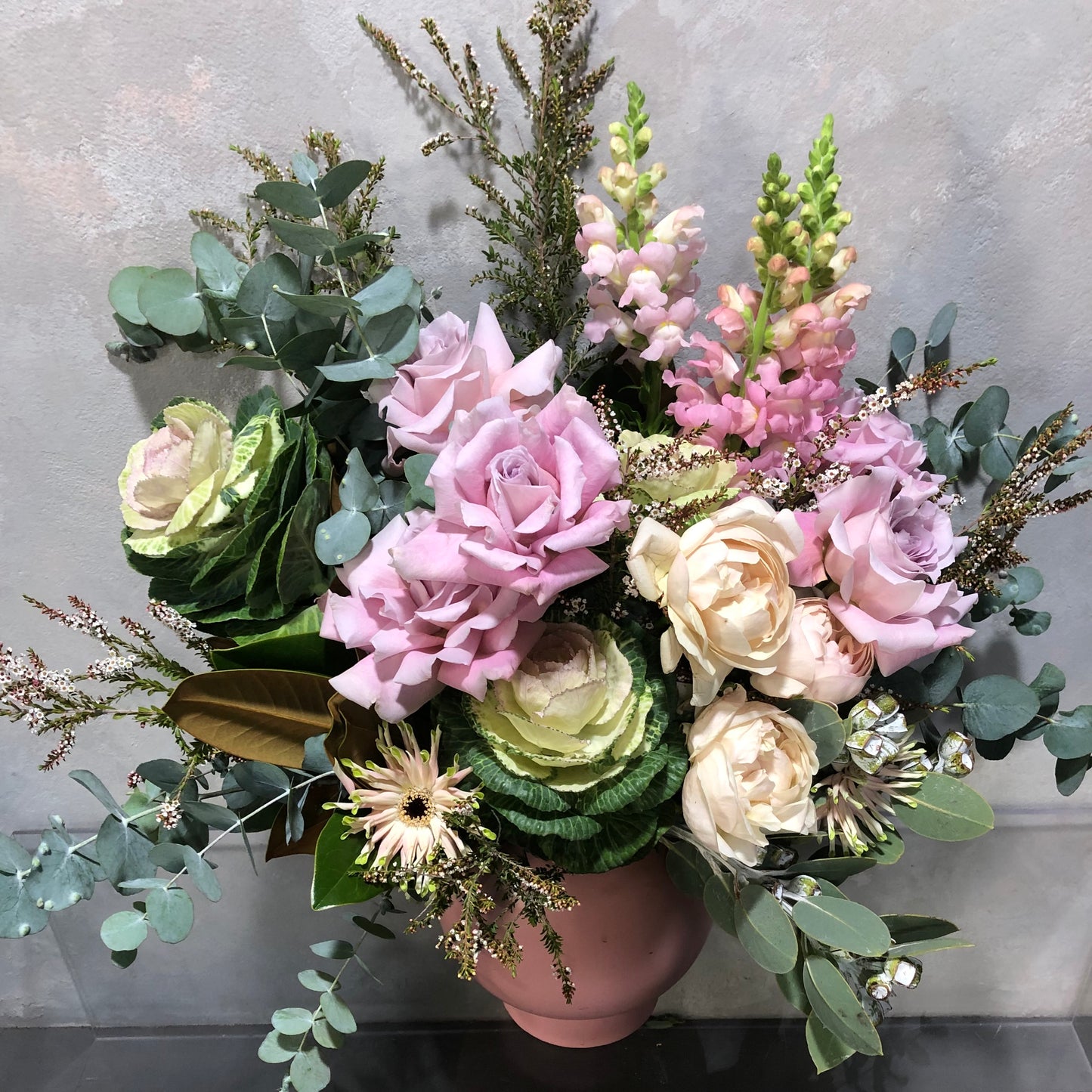 The Winter Garden Vase by Flowers on Norton St showcases a stunning bouquet of mixed flowers, featuring pink and white roses, eucalyptus leaves, pink snapdragons, ornamental kale, and native foliages, beautifully arranged against a neutral textured background.