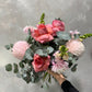 The "Pretty in Pink" bouquet from Flowers on Norton St beautifully presents an arrangement of pink roses, lush dahlias, and snapdragons nestled among eucalyptus leaves, all displayed against a textured gray backdrop.