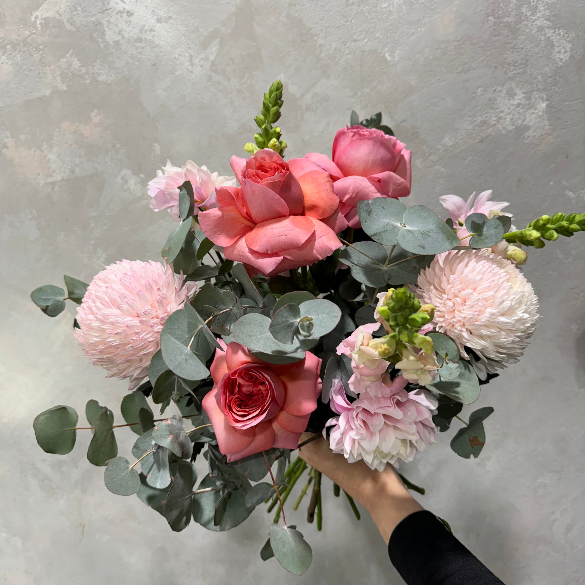 The "Pretty in Pink" bouquet from Flowers on Norton St beautifully presents an arrangement of pink roses, lush dahlias, and snapdragons nestled among eucalyptus leaves, all displayed against a textured gray backdrop.